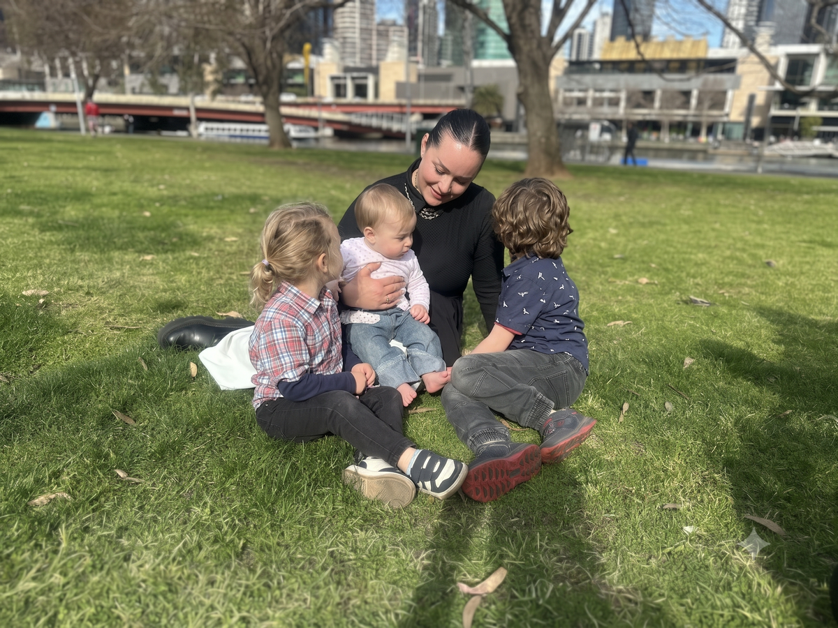 A woman sitting on a grassy park lawn with three children outdoors. The background shows trees, a bridge, and city buildings in soft focus.