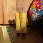 Two yellow beeswax taper candles standing on a dark wooden table, with a rustic wood plank wall behind them and a colorful floral fabric bag partially visible on the right.