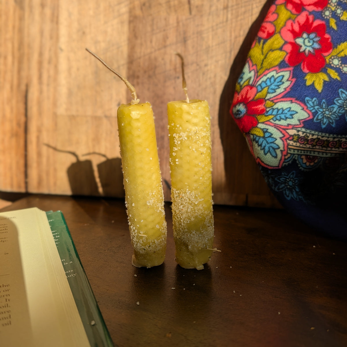 Two yellow beeswax taper candles standing on a dark wooden table, with a rustic wood plank wall behind them and a colorful floral fabric bag partially visible on the right.
