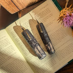 Two dark, textured candles with wax tips rest on an open vintage book, styled with dried flowers on a dark wooden table.