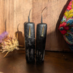 Two black pillar candles with a distressed, weathered finish and small gold-toned metal wicks, standing on a dark wooden surface against a rustic wood plank background.