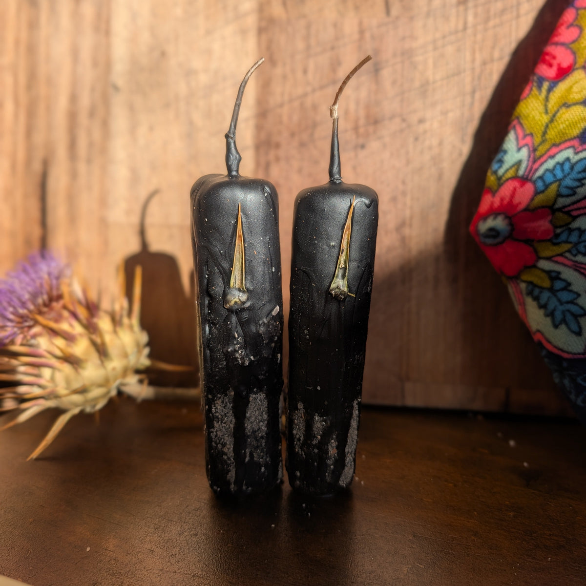 Two black pillar candles with a distressed, weathered finish and small gold-toned metal wicks, standing on a dark wooden surface against a rustic wood plank background.