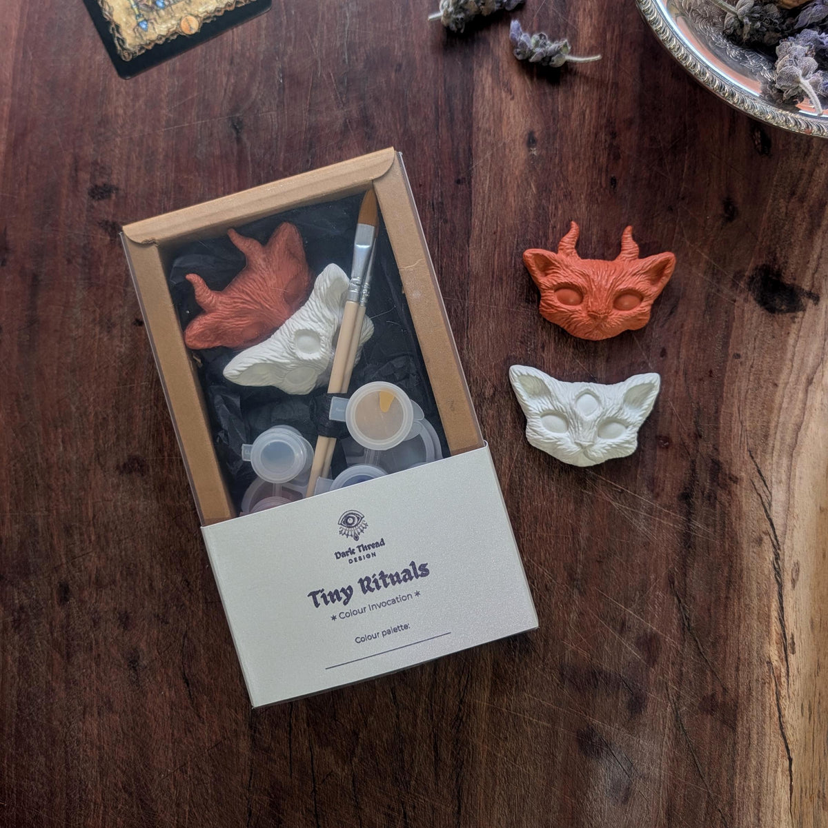 A boxed miniature painting kit labeled 'Tiny Rituals Colour Introduction 4' sits on a dark wooden table, with two painted clay animal faces placed beside the box.